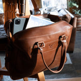 Tan leather briefcase with iPad and a magazine peeking out of the unzipped top, resting on a wooden chair indoors