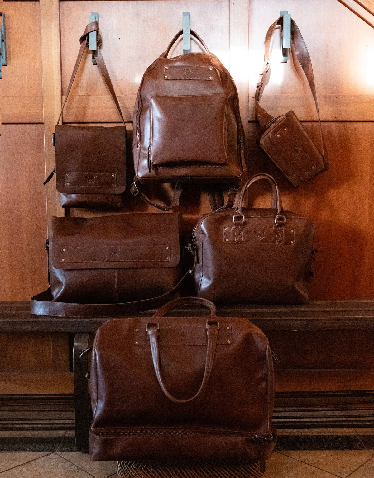 Lifestyle display of the full Will Leather Goods Field Collection in brown leather, including the messenger bag, backpack, belt bag, briefcase, and duffle hung on wall pegs and arranged on a bench.
