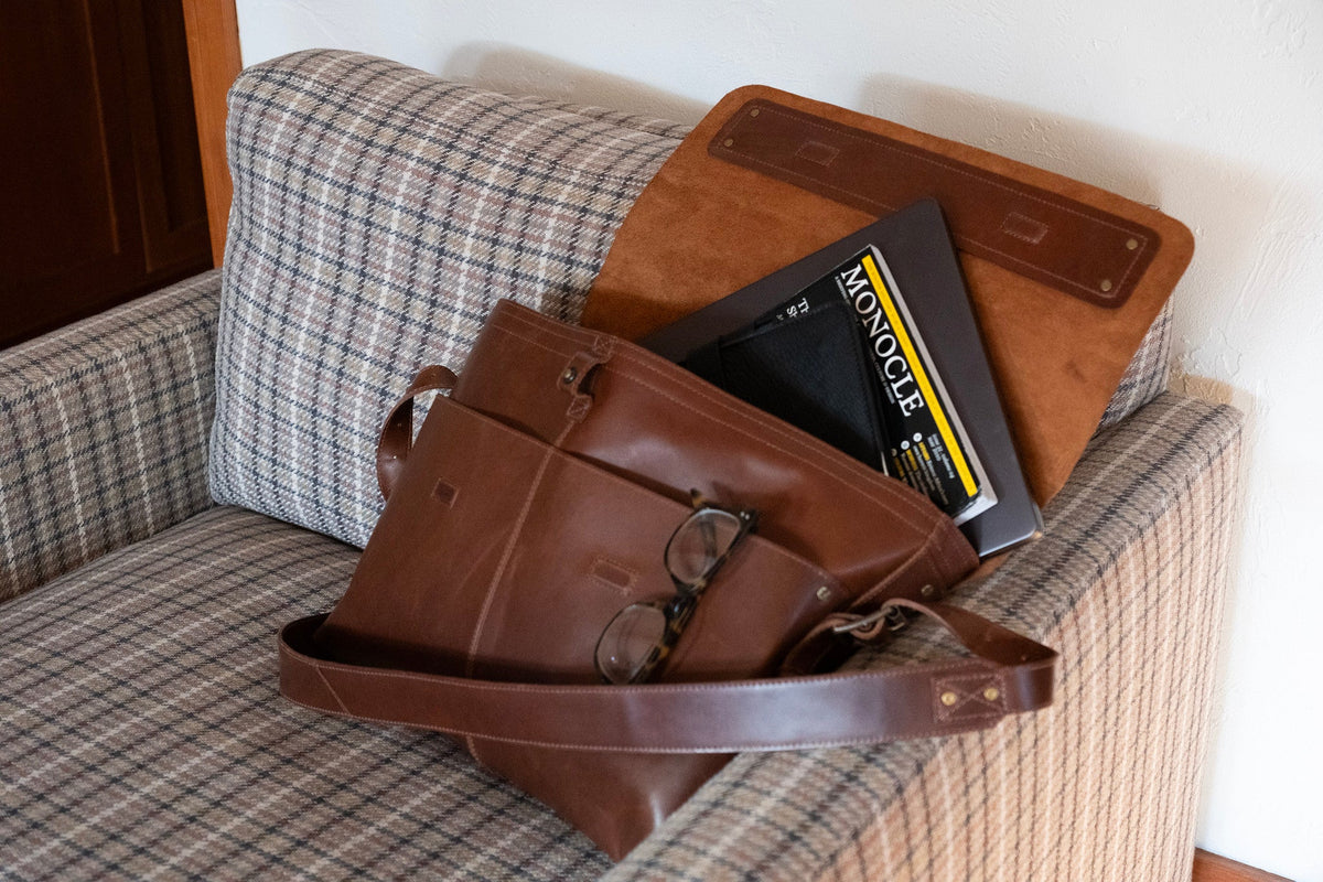 Lifestyle photo of the Will Leather Goods Field Messenger in tan leather resting on a plaid armchair next to a laptop, a Monocle magazine, glasses, and a leather portfolio.