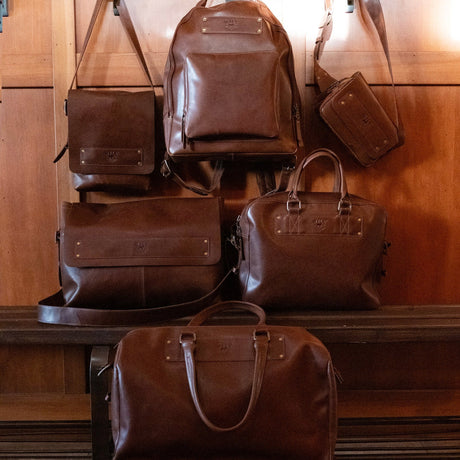 Collection display of WILL Leather Goods brown leather bags including a briefcase, backpack, messenger bag, duffel, and crossbody pouch on a wooden bench