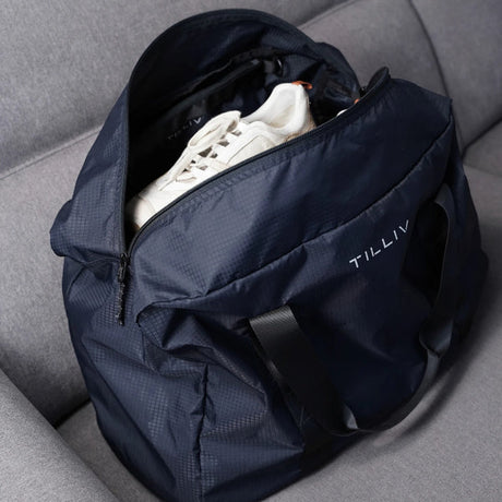 A navy TILLIV Pakke duffel slid over the extended handle of a silver rolling suitcase using its rear pass-through strap