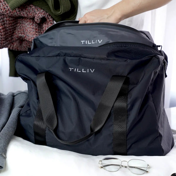 A hand lowering the grey Kamper vacuum pack into a navy TILLIV Pakke duffel on a bed, folded knitwear and glasses alongside
