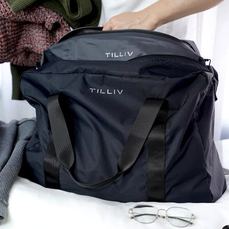 A hand lowering the grey Kamper vacuum pack into a navy TILLIV Pakke duffel on a bed, folded knitwear and glasses alongside