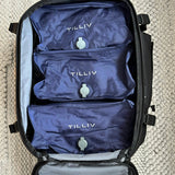 Three Midnight Blue TILLIV Stak packs filling the open main compartment of a travel case on a woven rug