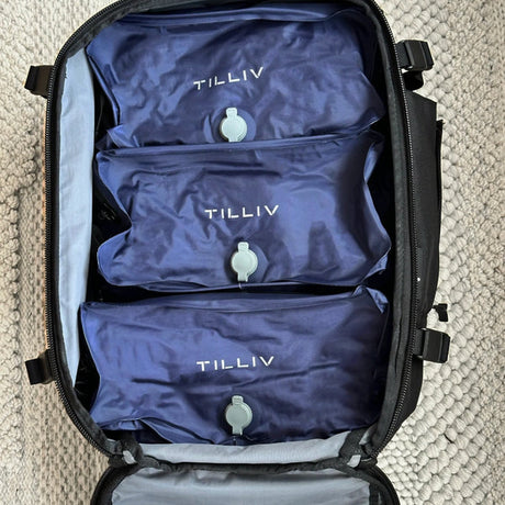 Three Midnight Blue TILLIV Stak packs filling the open main compartment of a travel case on a woven rug