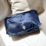 A packed Midnight Blue TILLIV Stak resting on a bed beside a cream cushion