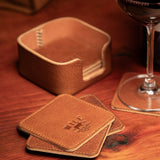 Lifestyle photo of the tan leather Set of Eight Coasters holder on a wood table beside a glass of red wine, with two coasters stacked in front.